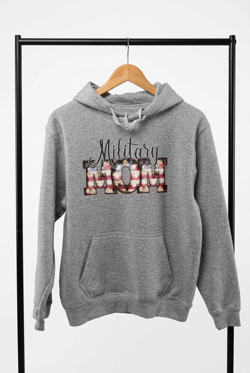 A heather gray pullover hoodie hanging on a wooden hanger against a black wire grid display wall. The hoodie features a front graphic that reads 'Military Mom' with 'MOM' filled with a red, white, and blue striped pattern resembling the US flag.
