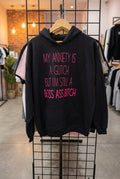 A black pullover hoodie hanging on a wire display rack, featuring the phrase 'MY ANXIETY IS A GLITCH. BUT I'M STILL A BOSS ASS BITCH' printed in bright pink text across the front.