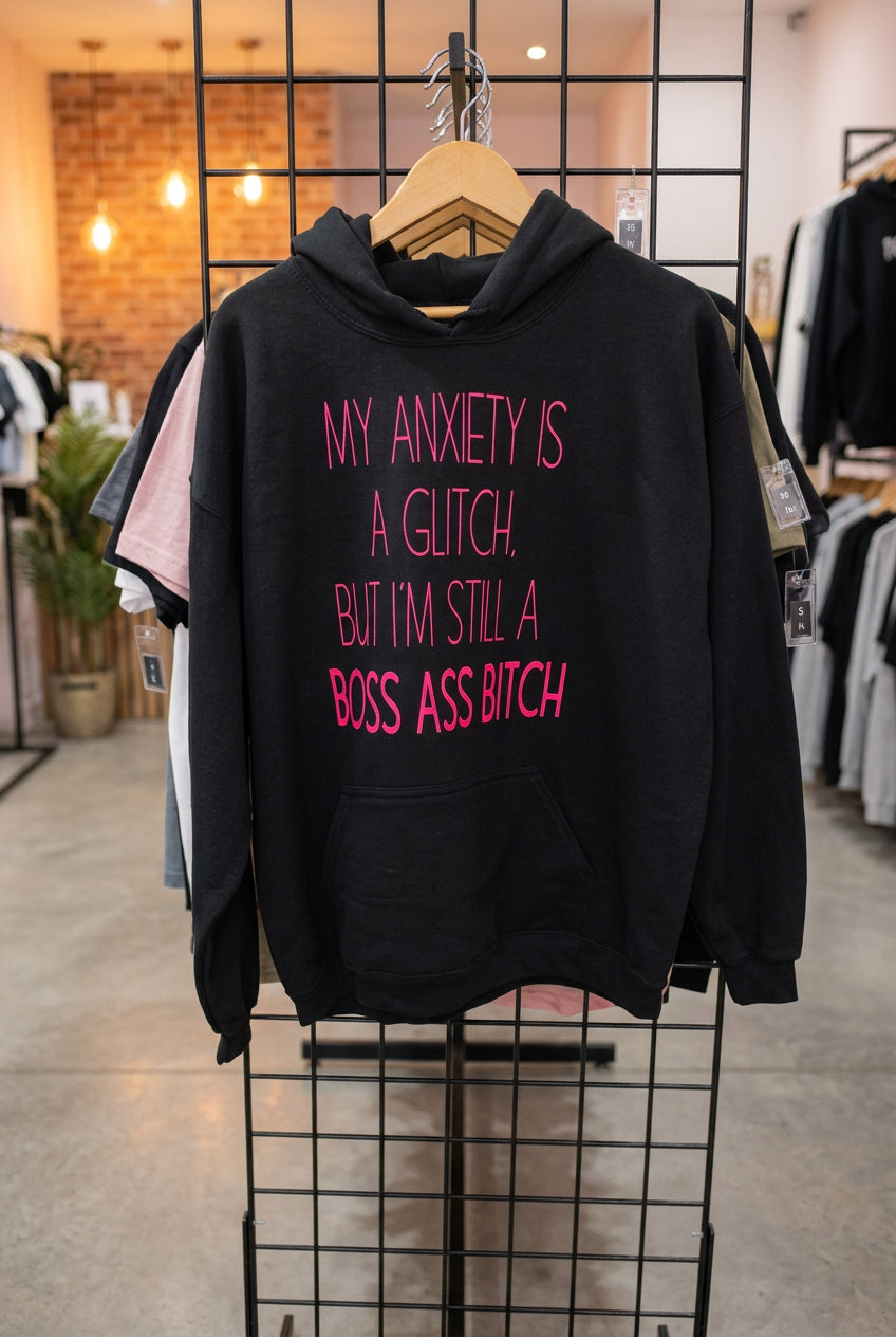 A black pullover hoodie hanging on a wire display rack, featuring the phrase 'MY ANXIETY IS A GLITCH. BUT I'M STILL A BOSS ASS BITCH' printed in bright pink text across the front.