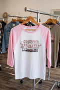 A pink and white raglan baseball t-shirt hanging on a wooden hanger against a black wire grid display wall. The shirt features a graphic print on the front with the text 'COUNTRY MAMA' and a longhorn illustration.