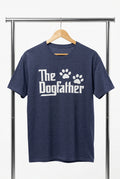 A navy blue heather crewneck t-shirt hanging on a wooden hanger against a black wire grid wall. The shirt features the text 'The Dogfather' in a white stylized font, with two white paw prints replacing the letter 'o' in 'Dog'.