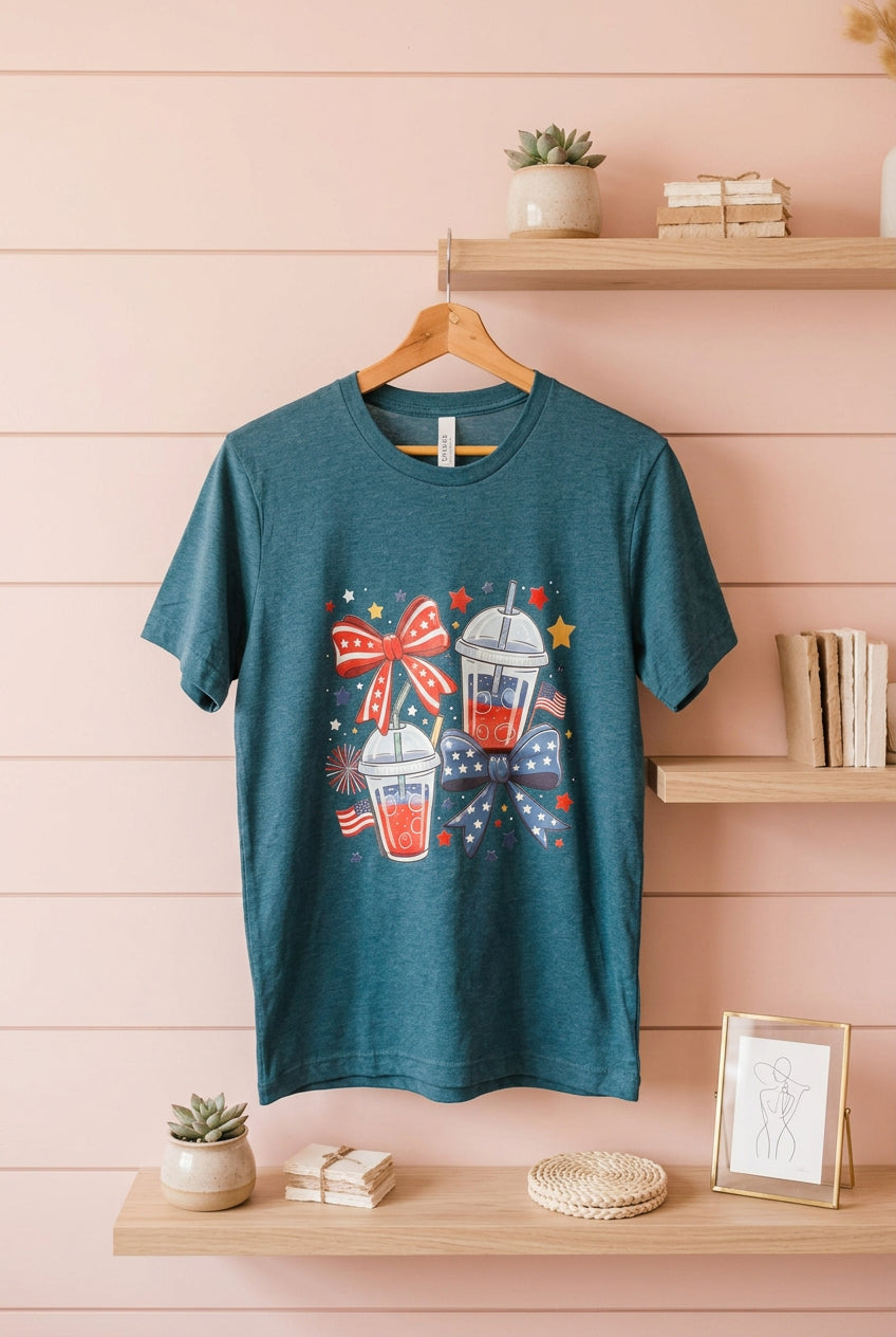 A teal heather short-sleeve t-shirt hanging on a wooden hanger against a pink wood-paneled wall, featuring a patriotic red, white, and blue graphic of two iced drinks with a bow and stars.