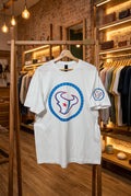 A white short-sleeve t-shirt hanging on a wooden hanger against a black wire grid wall. The shirt features a large circular graphic on the front with a blue segmented border, a stylized red and blue bull head logo, and a small matching logo on the left sleeve.