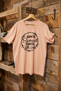 A light peach-pink short-sleeve t-shirt with a black graphic print reading 'SORTA Sweet SORTA beth Dutton' inside a thorny circle, hanging on a wooden hanger against a black wire grid display wall.