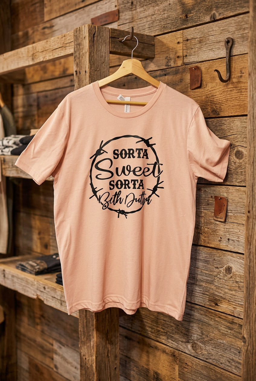 A light peach-pink short-sleeve t-shirt with a black graphic print reading 'SORTA Sweet SORTA beth Dutton' inside a thorny circle, hanging on a wooden hanger against a black wire grid display wall.