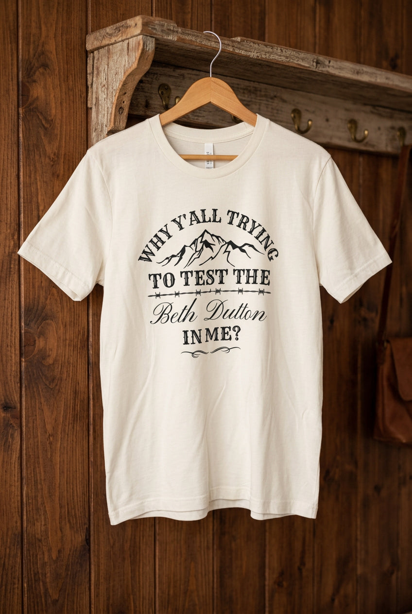 An off-white short-sleeve graphic t-shirt hanging on a wooden hanger against a black wire grid wall. The shirt features a black printed graphic with mountains and the text 'WHY Y'ALL TRYING TO TEST THE Both Dutton INME?'.