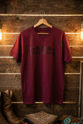 A short-sleeve crewneck t-shirt in heathered purple, hanging on a wooden hanger against a black wire grid wall. The front of the shirt features the text 'Beth Dutton STATE OF MIND' printed in black.