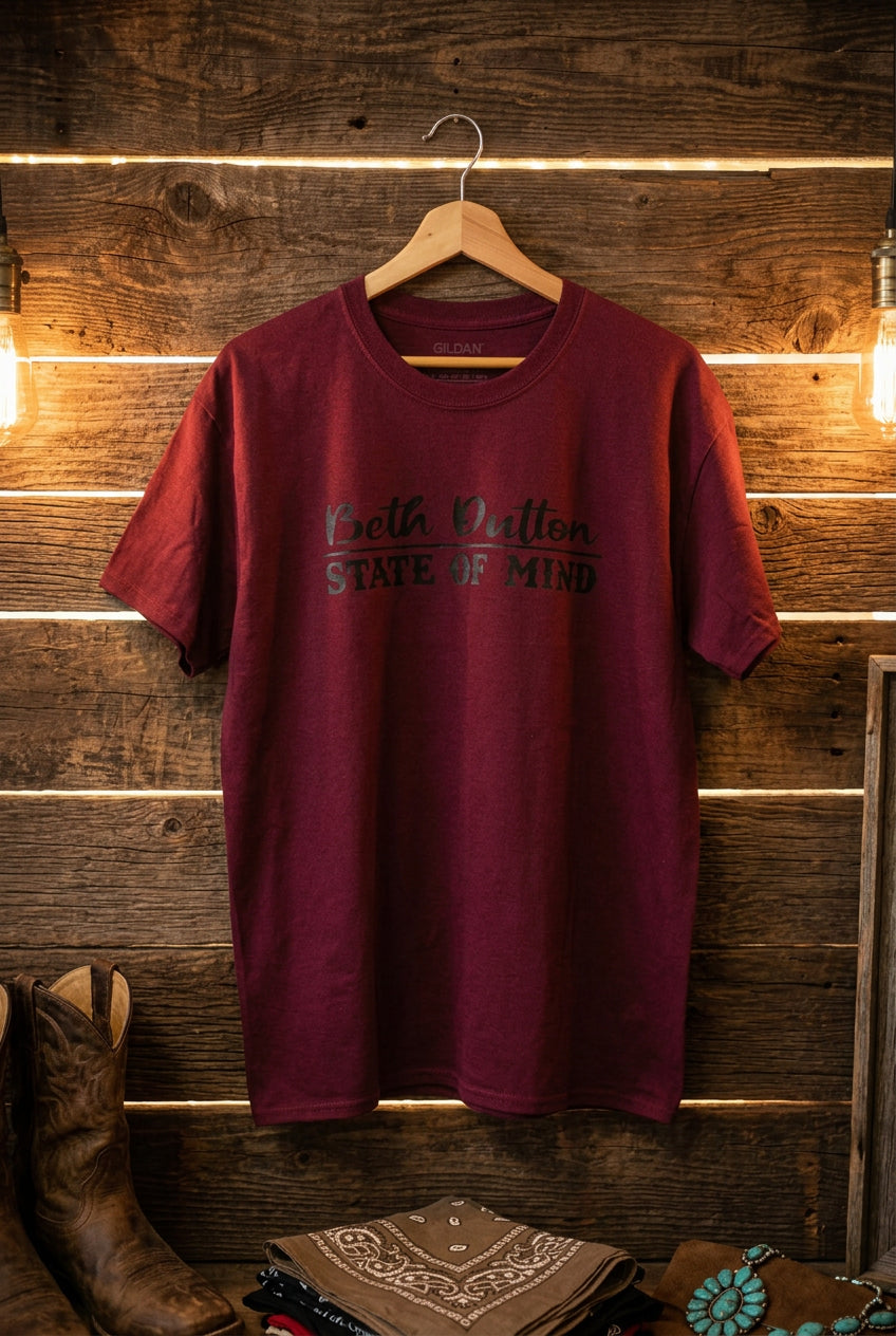 A short-sleeve crewneck t-shirt in heathered purple, hanging on a wooden hanger against a black wire grid wall. The front of the shirt features the text 'Beth Dutton STATE OF MIND' printed in black.