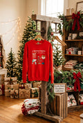 A red long-sleeve crewneck sweatshirt featuring a ' Christmas' graphic print with a boy and his dog, displayed on a white mannequin with denim shorts against a white wood-paneled background.