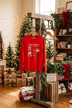 A red long-sleeve crewneck sweatshirt featuring a ' Christmas' graphic print with a boy and his dog, displayed on a white mannequin with denim shorts against a white wood-paneled background.
