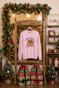 A light pink crewneck sweatshirt hanging on a wooden hanger against a wall backdrop. The sweatshirt features a graphic print of a taza de cafecito with the words 'BUT FIRST Cafecito' in stylized lettering.