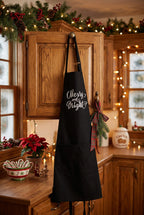 A black kitchen apron with 'Merry and Bright' written in white script hangs on a wooden cabinet in a cozy, warmly lit holiday kitchen decorated with garlands, lights, and poinsettias.