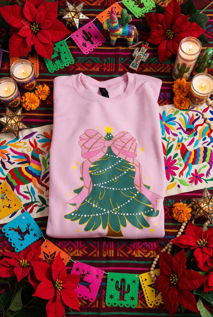 A folded pink crewneck sweatshirt featuring a graphic of a Christmas tree decorated with a large pink concha bow, white lights, and gold outlines.