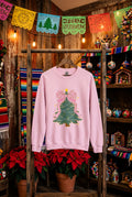A pink crewneck sweatshirt displayed on a wooden rack. The sweatshirt features a graphic of a Christmas tree decorated with a large pink concha bow and gold star details.