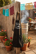 A black apron with white Spanish text and a bee graphic, hanging on a knob at an outdoor bbq.