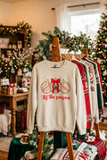 A sand colored crewneck sweatshirt displayed on a wood rack against sweaters. The sweatshirt features a graphic of three talavera decorated Christmas ornaments in brown, and white, with the red cursive text 'tis the season' printed below.