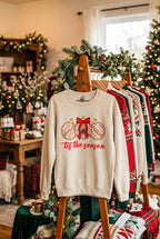 A sand colored crewneck sweatshirt displayed on a wood rack against sweaters. The sweatshirt features a graphic of three talavera decorated Christmas ornaments in brown, and white, with the red cursive text 'tis the season' printed below.