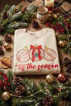 A folded beige crewneck sweatshirt featuring a graphic of three decorated Christmas ornaments in brown, red, and white, with the phrase 'tis the season' printed in red cursive text below.