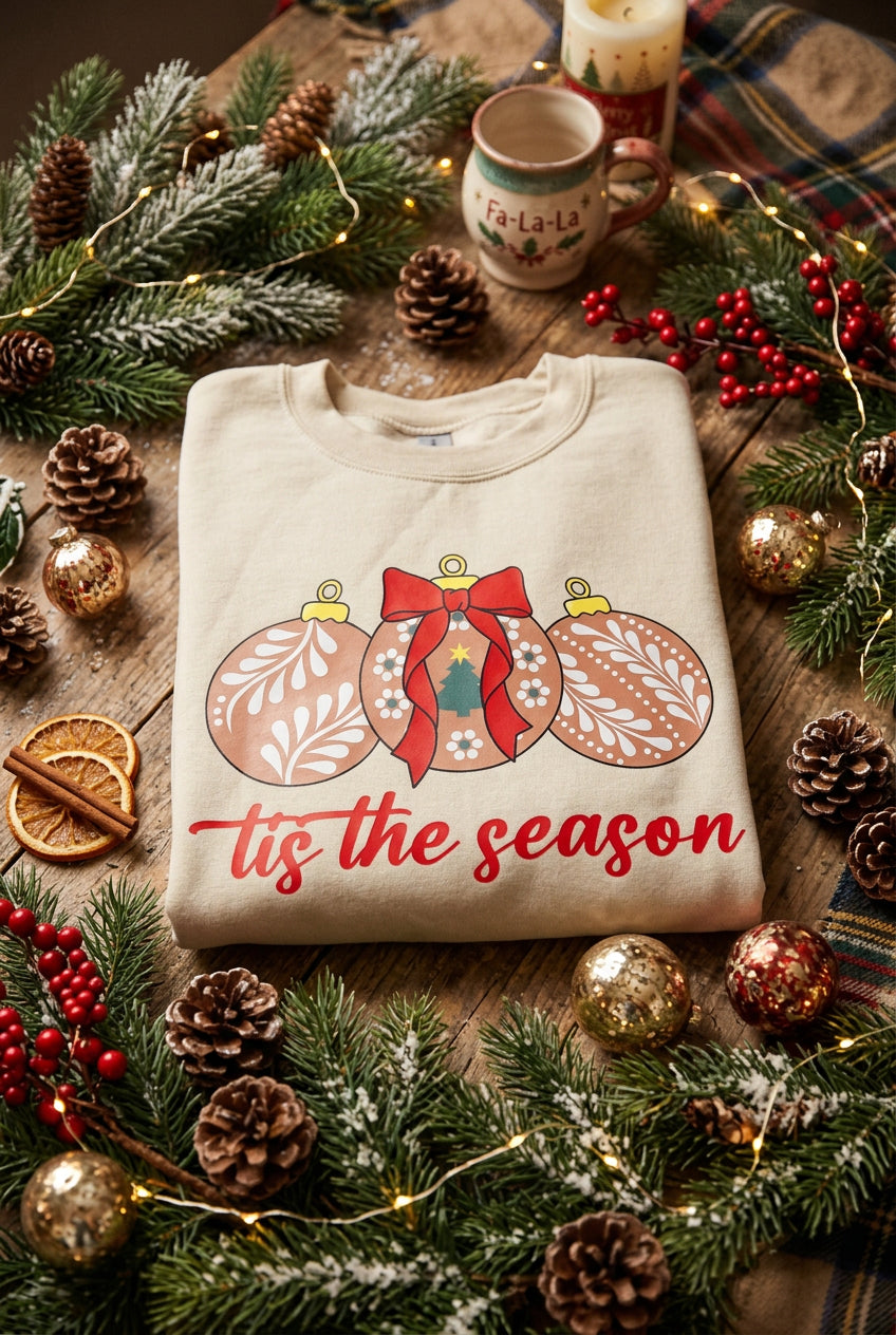 A folded beige crewneck sweatshirt featuring a graphic of three decorated Christmas ornaments in brown, red, and white, with the phrase 'tis the season' printed in red cursive text below.