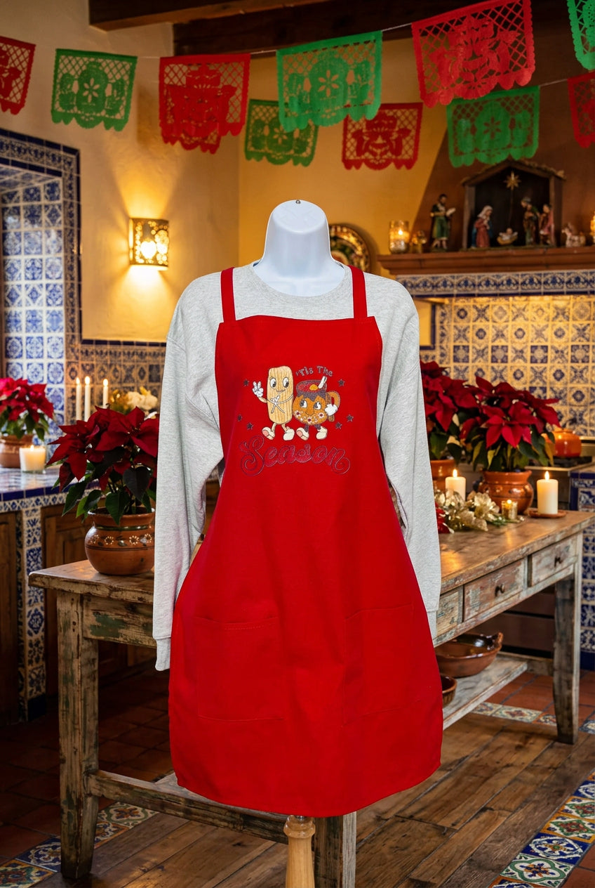 A red apron with a cartoon tamale walking with a tazita de ponche graphic and the text 'tis the season' displayed on a mannequin, set in a decorated holiday kitchen or cafe interior with poinsettias, candles, and papel picado banners.