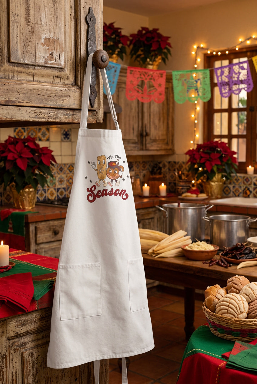 A white apron with a cartoon graphic of a tamale walking with a tazita de ponche, with the text 'Tis the Season' in red script. The apron is displayed on a knob in a kitchen.