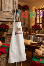 A white apron with a cartoon graphic of a tamale walking with a tazita de ponche, with the text 'Tis the Season' in red script. The apron is displayed on a knob in a kitchen.